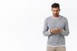 © Cookie Studio - Young modern man in grey sweater using smartphone with busy serious expression, wear wireless earphones, holding mobile phone, typing message, chatting about business with coworkers in messanger