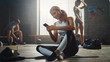 © Gorodenkoff - Happy and Smiling Beautiful Athletic Young Woman is Using a Smartphone while Sitting on a Floor in a Loft Gym. She's Typing a Message and Smiling. A Man Exercises in the Background.