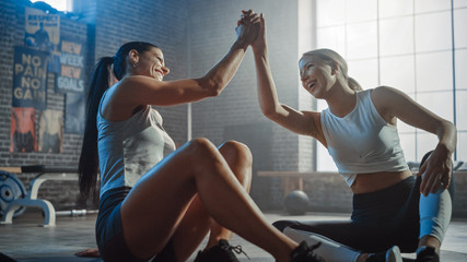 Poster - Two Beautiful Fit Athletic Girls Sit on a Floor of Industrial Loft Gym. They're Happy with their Training Program and Successfully Give a High Five.