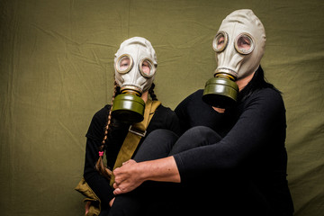 A woman and a little girl in black clothes and gas masks sit in the shelter. Mother and daughter in chemical protection products sit on a green background. Family quarantine concept.