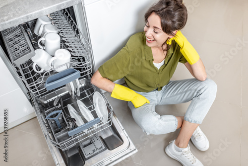 Portrait of a happy housewife sitting near the dishwasher with clean dishes on the kitchen at home. Easy house work with kitchen appliances concept