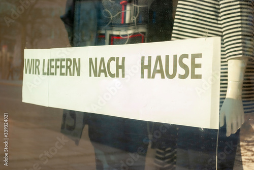 Schaufenster von Ladengeschäft mit Schild „Wir liefern nach Hause“, in Deutsch, mit Auslagen und die Spiegelung der Umgebung in Unschärfe zur Zeit der Corona Covid-19 Pandemie im März 2020
