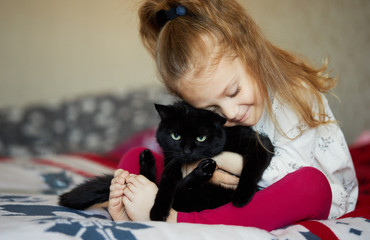 Portrait of a little cute child girl who hugs a black cat with tenderness and love and smiles with happiness