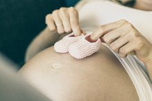 Expectant Mother Applying Pink Baby Shoes To Big Naked Belly. Pregnant Woman Enjoying Expecting Daughter. Anticipation Concept