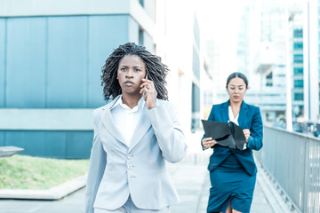 Focused young female professional talking on cell on her way to office. Young African American business woman walking outside in city. Communication concept