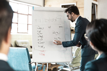 Confident bearded speaker talking and writing on whiteboard. Group of employees looking at presentation of new project at briefing. Business meeting concept