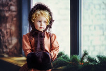 Little Beautiful 4 Years Old Curly Blonde Girl In Brown Vintage Retro Hat And Dress Sit On Window Sill With Hands In Hand Warmer. Vintage Post Card With Child. Daughter Waiting For Parents