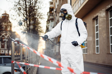 A man in protective equipment disinfects with a sprayer in the city. Surface treatment due to coronavirus covid-19 disease. A man in a white suit disinfects the street and rails. Virus pandemic.