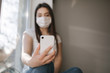 © Aleksandr - Attractive woman in medical mask sits on windowsill at home. Female in white t'shirt and white medical mask use phone