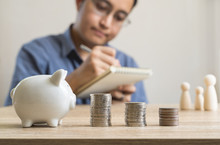 Money Savings Concepts  Piggy Bank And Stack Coins With Blur Man Who's Taking Notes About Saving Money For Family And Family Wooden Dolls.on Wooden Table With Blur Background And Copy Space.