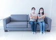 © peenat - Asian women wearing masks sit together on sofa without space and to cross sign for social distancing to reduce spreading of coronavirus. Work from home, social distancing and covid-19 concept.