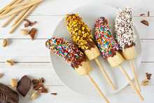 Frozen Chocolate Dipped Banana Pops With Sprinkles, Nuts And Coconut. Overhead View Table Scene On A Wood Background.