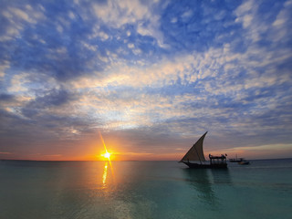 Wall Mural - Calm indian ocean in the rays of the setting sun and blue sky with small clouds. a boat with a sail goes on the ocean.