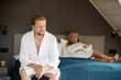 © Comeback Images - Depressed middle aged man in white bathrobe sitting on edge of bed being deep in bad thoughts, his dissatisfied wife or lover lying in background