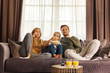 © Comeback Images - Front view of captivated family of mother, father and little daughter watching movie and eating popcorn sitting on sofa at home