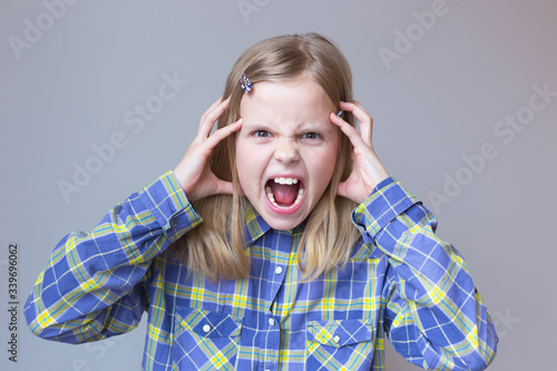 White girl blonde 10 years old is very angry. She grabbed her head in her hands. Portrait on a gray background. Emotion of aggression and anger. Help me. Leave me alone.Selective focus.checkered shirt