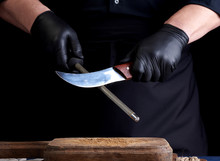 Chef In A Black Shirt And Black Latex Gloves Sharpen A Kitchen Knife On An Iron Sharpener With A Handle