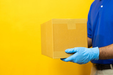 Delivery man blue uniform wearing rubber gloves holding parcel cardboard box on yellow background.