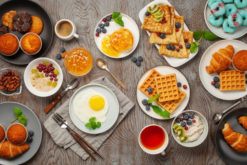 Wall Mural - Continental breakfast captured from above (top view, flat lay). Coffee, tea, croissants, jam, egg, pancakes, maffins and oatmeal. Wooden background. Family breakfast table.