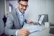 © Dusan Petkovic - Attractive smiling caucasian classy unshaven businessman in suit and with eyeglasses looking at paperwork while sitting in office. On table are mug with coffee, laptop and paperwork.