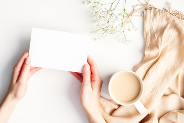 Female hands holding blank paper card over cozy home white desk with cup of coffee, blanket, flowers. Hygge, autumn fall, comfort concept. Flat lay, top view, copy space