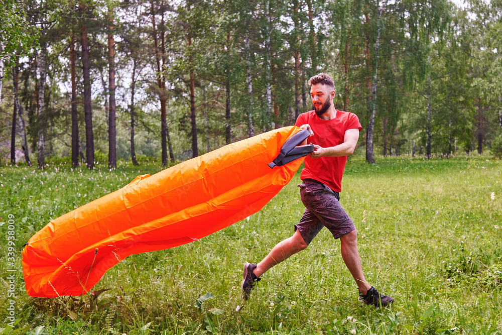 A young man inflates a lamzac with air. A man inflates an air sofa. on the background of the forest - obrazy, fototapety, plakaty 
