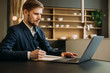 © Girts - Confident businessman working remotely from a home kitchen with a laptop and notebook. Side view of a young man in smart casual wear making notes during zoom video meeting conference call remote