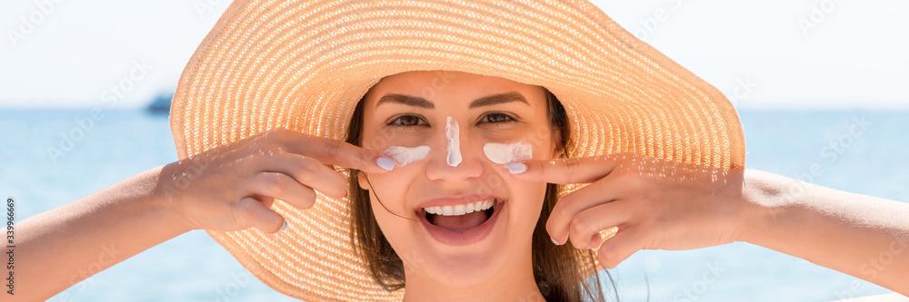 Beautiful young woman in hat is applying sunblock under her eyes and on her nose like Indian. Sun protection concept - obrazy, fototapety, plakaty 