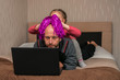 A child puts a bright wig on the head of a bald man. A little girl prevents dad from working at a computer. telecommuting in quarantine. A fun family event.