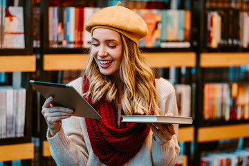 Portrait of attractive blonde woman posing next to shelves with books at book store. Tablet and book decision. 