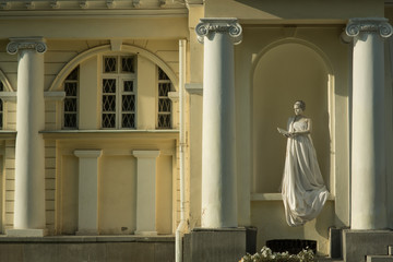 Wall Mural - Live sculpture of a young woman pregnant in a long white dress with a book, full-length, against the background of a yellow building with white columns