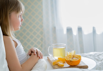 Little sick girl in bed with cup of antipyretic drugs for colds,flu.Tea with citrus vitamin C,ginger root,lemon,orange.Wooden tray. Home self-treatment.Medical quarantine covid-19 coronavirus therapy