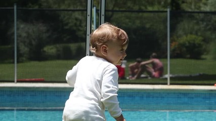 Wall Mural - Happy baby standing by pool leaning on fence prevention, toddler infant learning to stand smiling