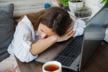 Caucasian teenager schoolgirl taking a nap with laptop computer and cup of tea on wooden table. Difficult, debilitating distance learning during the quarantine period coronavirus. Exam preparation.