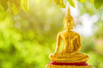 A golden Buddha statue sitting on a golden base with green blurred nature background with green leaves nature foreground, Buddhist Sabbath Day concept.