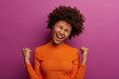 © WHstudio Leushin N - Hopeful optimistic dark skinned girl makes fist bump, cheers over something, gestures actively, happy with good luck, tilts head, dressed in orange jumper, isolated on purple wall, receives great news