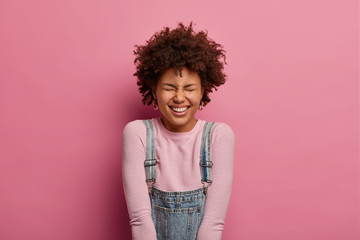 Beautiful carefree woman with toothy smile, laughs at something positive, expresses happiness, giggles over funny joke, wears turtleneck and overalls, makes happy reaction on something really good