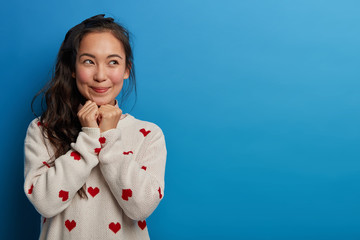 Wall Mural - Pretty young woman sees something wonderful, tempting to buy, keeps hands under chin, looks with smile aside, remembers pleasant moment, dressed in casual jumper, poses against blue background