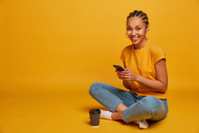 Photo Of Relaxed Dark Skinned Girl Happy To Receive Message From Lover, Holds Mobile Phone, Watches Funny Cute Video, Sits In Lotus Pose, Drinks Takeaway Coffee, Isolated On Yellow Background