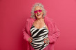 Horizontal shot of happy retired woman with grey curly hair smiles broadly, has white perfect teeth, wears bathing suit and coat, glad to meet with grandchildren, isolated on pink background