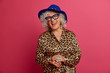 Horizontal shot of happy elderly woman happy to meet old friend, remembers her youth, wears elegant leopard dress and hat, being on pension, isolated over pink background. People and age concept