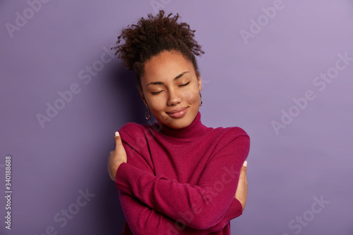 I love myself. Tender romantic woman embraces own body, hugs herself, closes eyes from pleasure, wears soft turtleneck just for cold weather, feels comfort, stands indoor against purple wall
