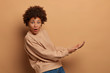 © WHstudio Leushin N - Isolated shot of surprised young Afro American woman pretends catching something big, keeps palms, holds invisible object from under, dressed in casual sweatshirt, isolated on beige background.