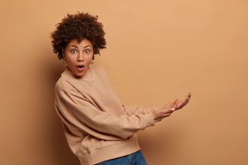 Isolated shot of surprised young Afro American woman pretends catching something big, keeps palms, holds invisible object from under, dressed in casual sweatshirt, isolated on beige background.