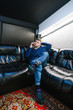 © bodnarphoto - Stylish young man sitting on a black leather sofa and talking on the phone. Trendy guy in jeans and trendy sneakers makes a phone call and relaxes on the sofa indoors. Vertical