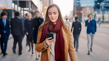 Wall Mural - Young Smart Casually Dressed Female is Using a Smartphone on a Street. Business People and Office Managers Walk Pass on Their Way to Work. She Looks Confident while Checking Her Cell and Walking.