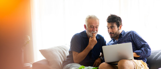 Wall Mural - Father and son family time together at home concept. Smiling old father and happy son sitting on sofa using digital laptop computer in living room at home