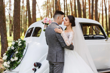 Elegant newlyweds, groom in grey suit and bride in wedding dress with long sleeves and long veil looking at each other near white Just Married car