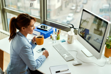 Wall Mural - Happy businesswoman drinking coffee while working on desktop PC in the office.