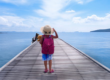 Bright Holiday Air, Wooden Bridge By The Sea, The Left-hander Carries A Doll In A Blue Suit And A Satchel In A Red Suitcase, Standing On A Wooden Bridge Standing On The Beach And Wearing A Sun Hat.